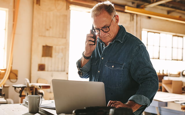 Älterer Herr steht telefonierend in einer Werkstatt und arbeitet an einem Laptop Älterer Herr steht telefonierend in einer Werkstatt und arbeitet an einem Laptop